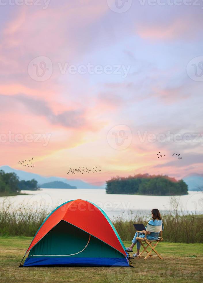 A sense of peace and serenity photo of female traveler sitting beside camping tent and using notebook laptop computer working from the lake side. There are group of birds flying in colorful sky