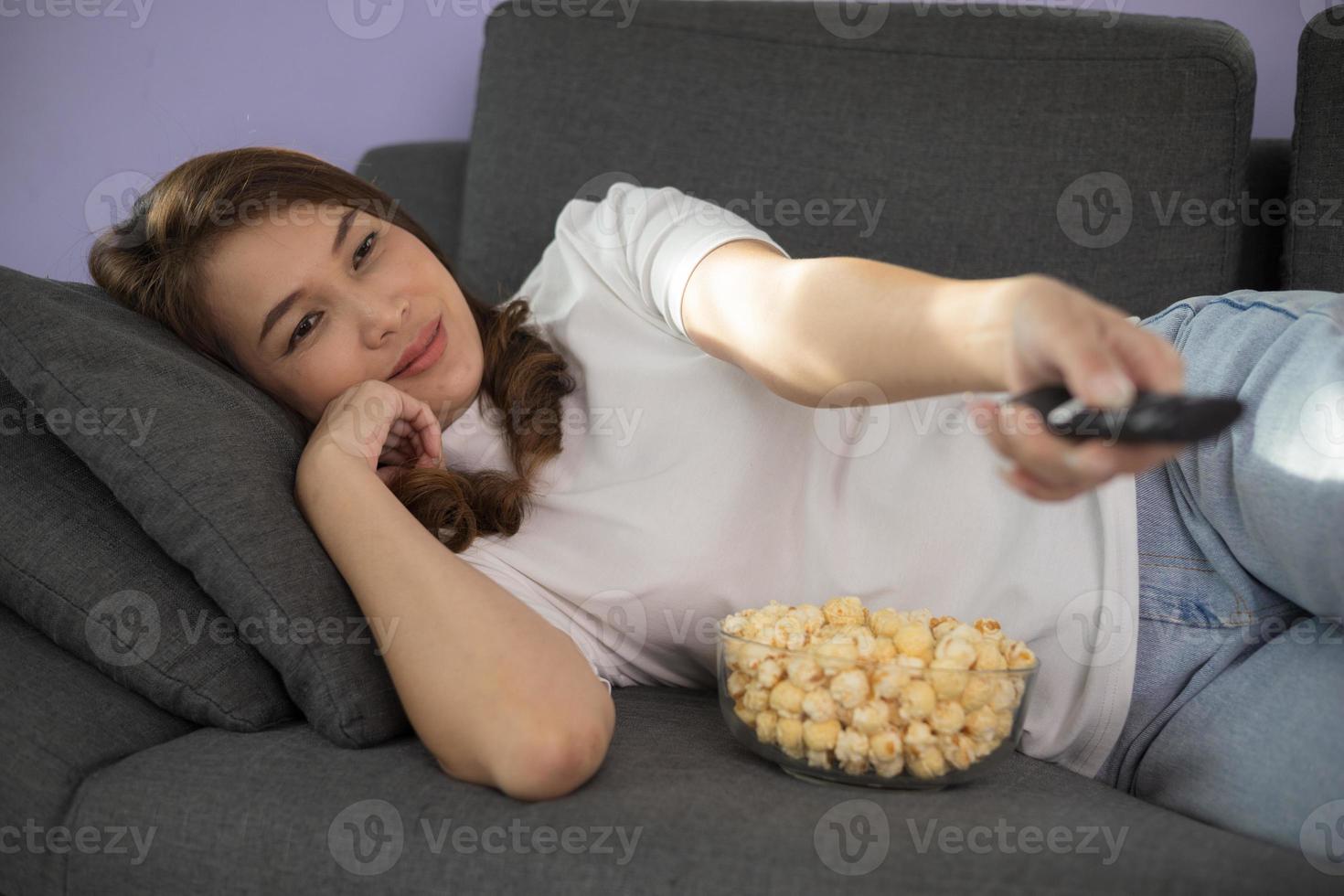 Beautiful and cute Asian woman reclining on sofa in living room and holding tv remote control with bowl of popcorn and sunlight shine from windows photo