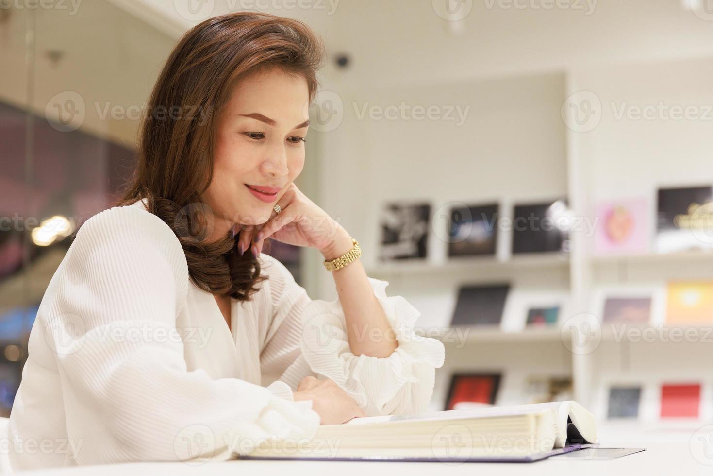 Portrait of Asain pretty cheerful female cozy sitting and reading book in library and leaning on glass wall with happy, focus and concentrate on contents photo