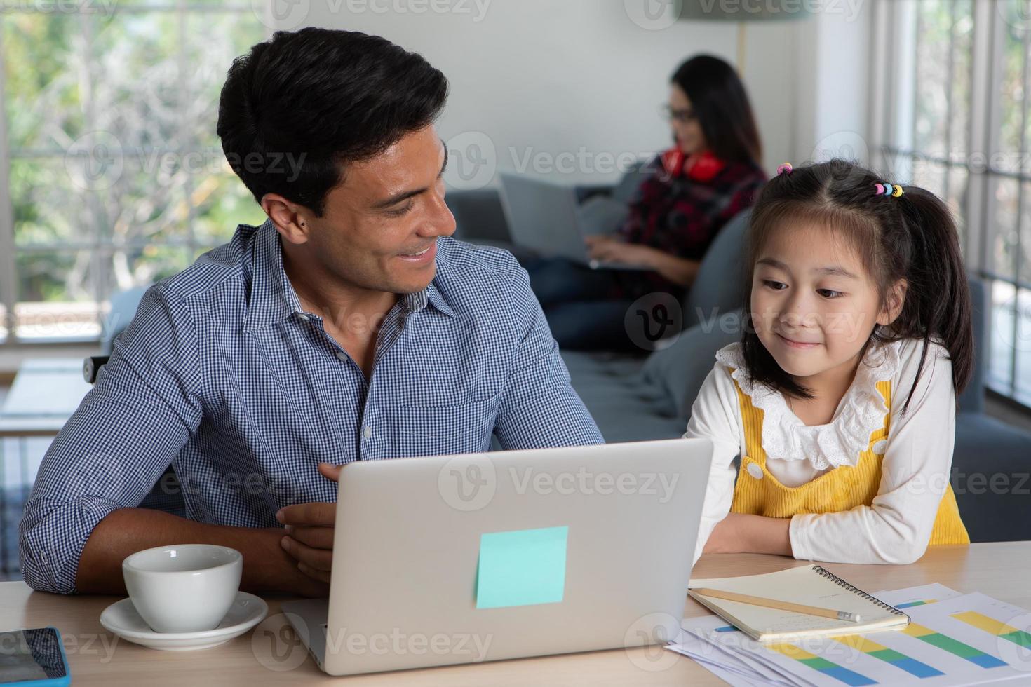 Mixed race family staying together, Caucasian father sitting and working at desk and teaching half-race little cute daughter white Asian mother using laptop computer on sofa. Idea for work at home photo