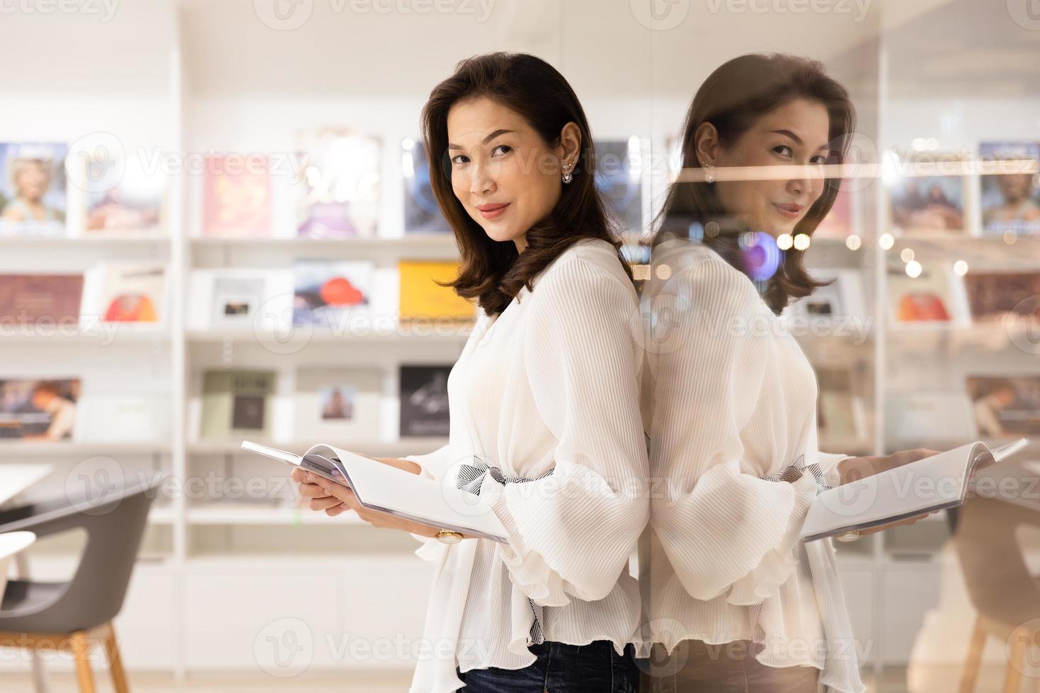 Portrait of Asain pretty cheerful female standing and reading book in library and leaning on glass wall with happy, focus and concentrate with reflection in clear mirror looking at camera photo