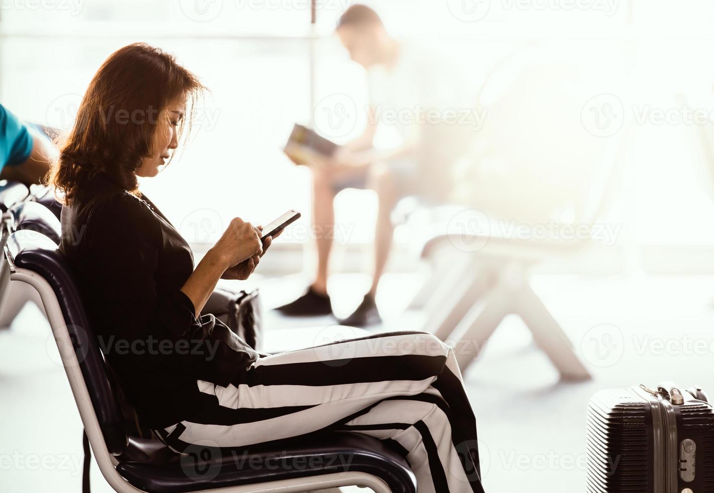 Woman sitting in airport lobby using smartphone and looking at screen while waiting for transit. Sunlight shining from outside photo