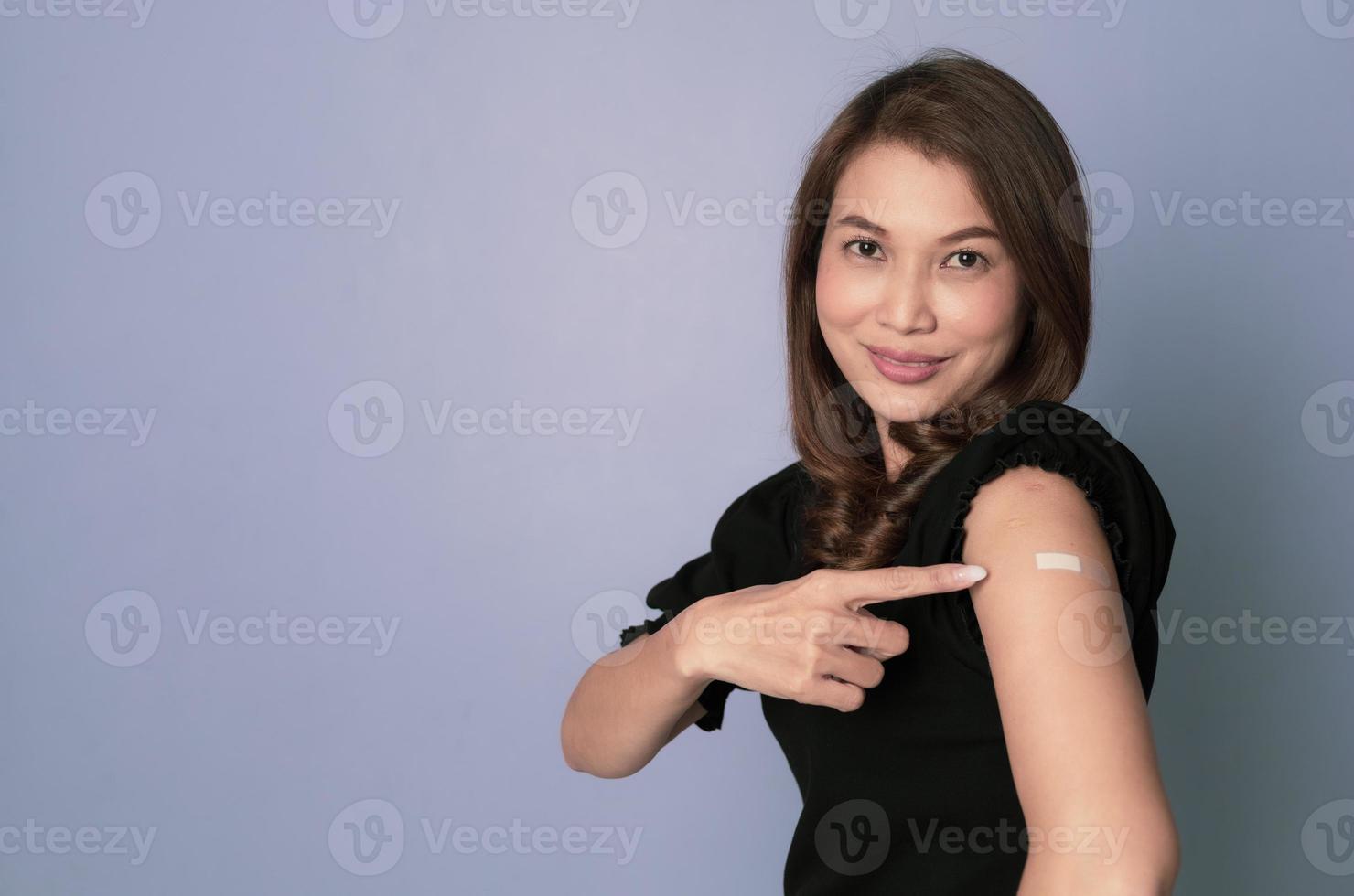 Asian woman sitting and using finger pointing to plaster on arm after getting covid-19 vaccine injection and looking to camera with confident photo