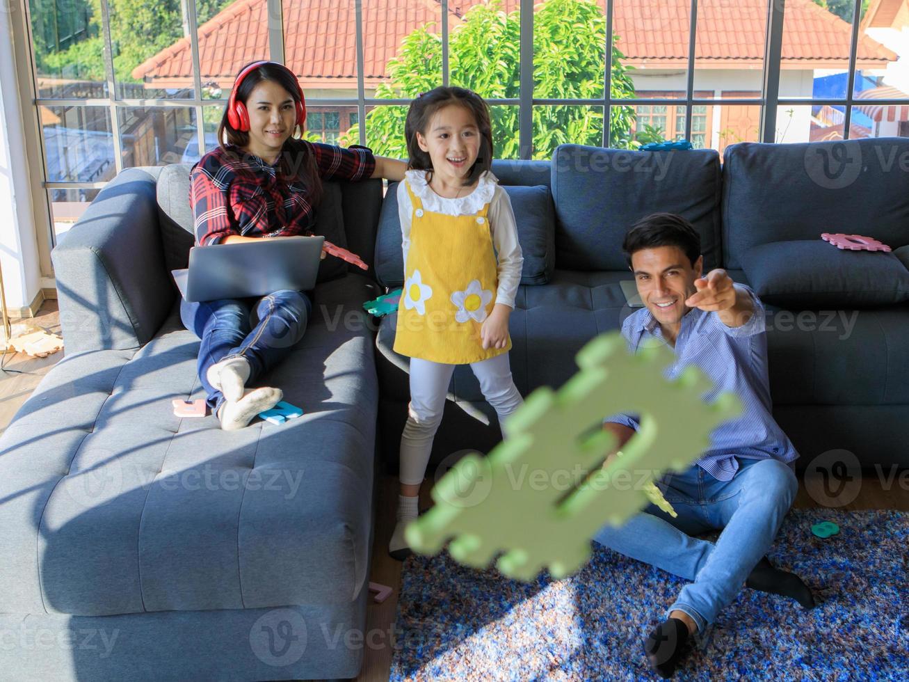 Three members of mixed races family, father, mother and kid daughter live together in home living room. Girl throwing toy with dad while mom working with notebook computer. Idea for work at home photo