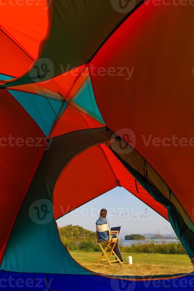 A woman sitting outside a camping tent and using laptop notebook computer working beside wide and big lake with clear blue sky and bids flying in natural scene wiith coffee cup. Taken from tent inside photo