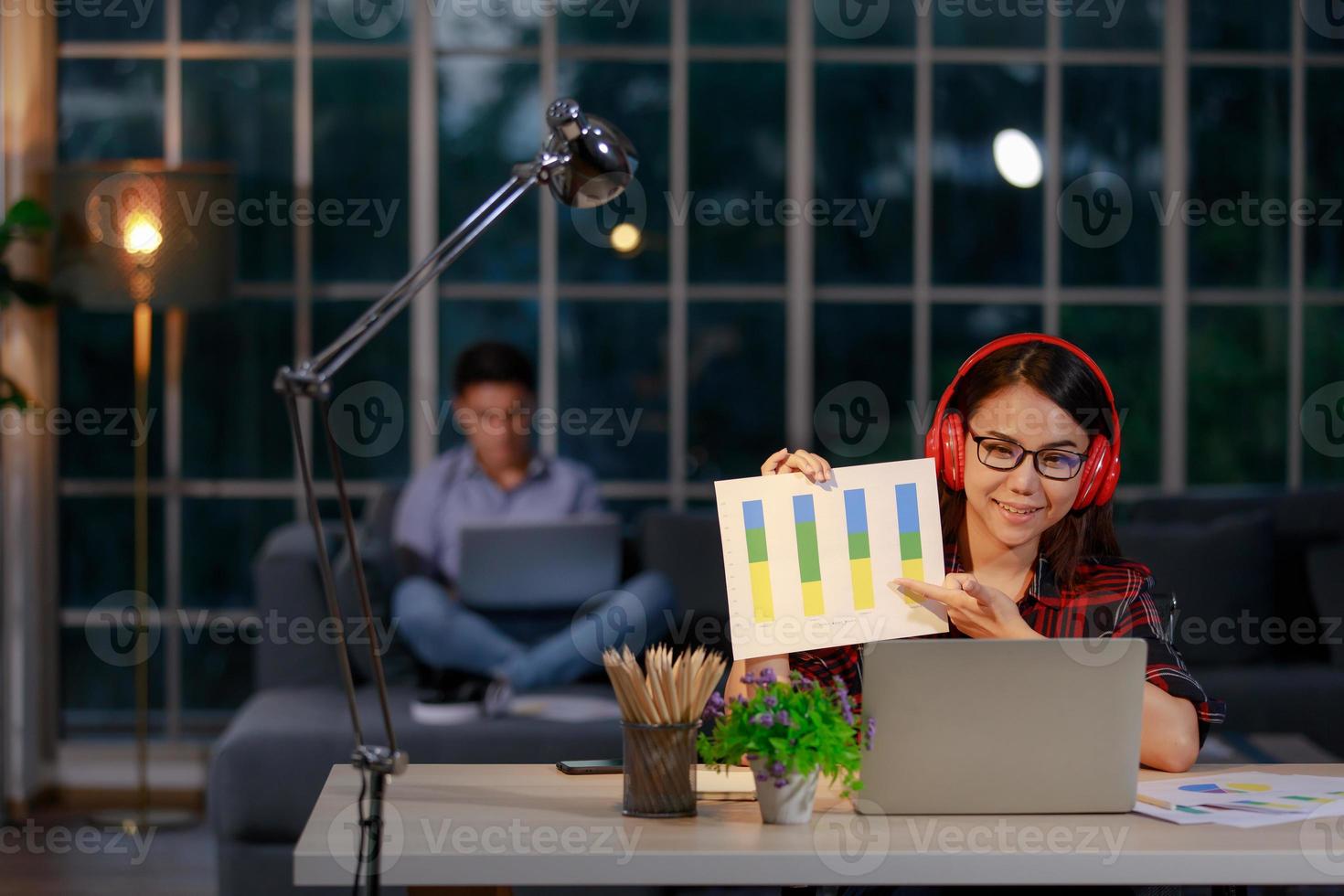 Man and woman couple sitting in living room at twilight in light from lamp and working on notebook computer, female presenting graph and chart to team or customer via laptop. Work at home concept photo