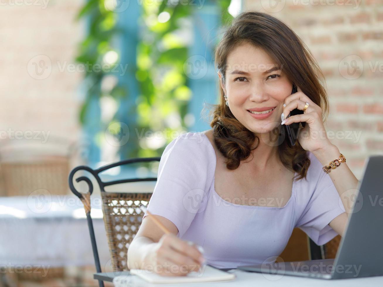 The beautiful middle-aged woman sitting in food shop and working with a laptop notebook computer and talking on the phone. Work from home and freelancer lifestyle concept photo