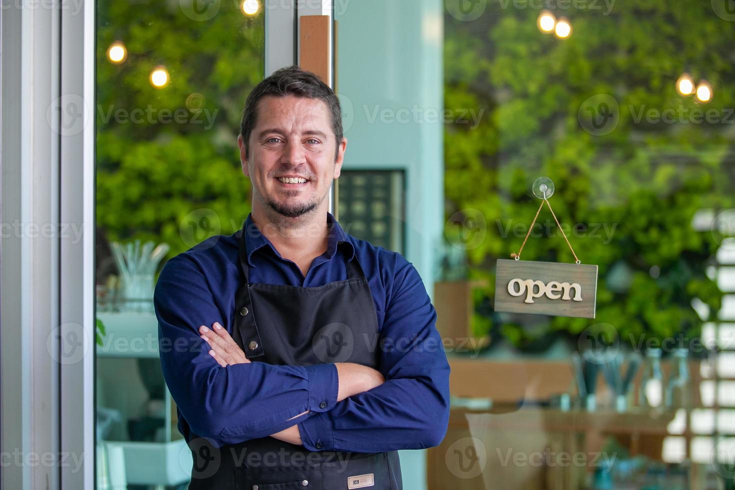 retrato de un dueño sonriente parado en la puerta de su café con un letrero abierto. dueño del café parado frente a la cafetería para dar la bienvenida al cliente y abrir la cafetería por la mañana. foto