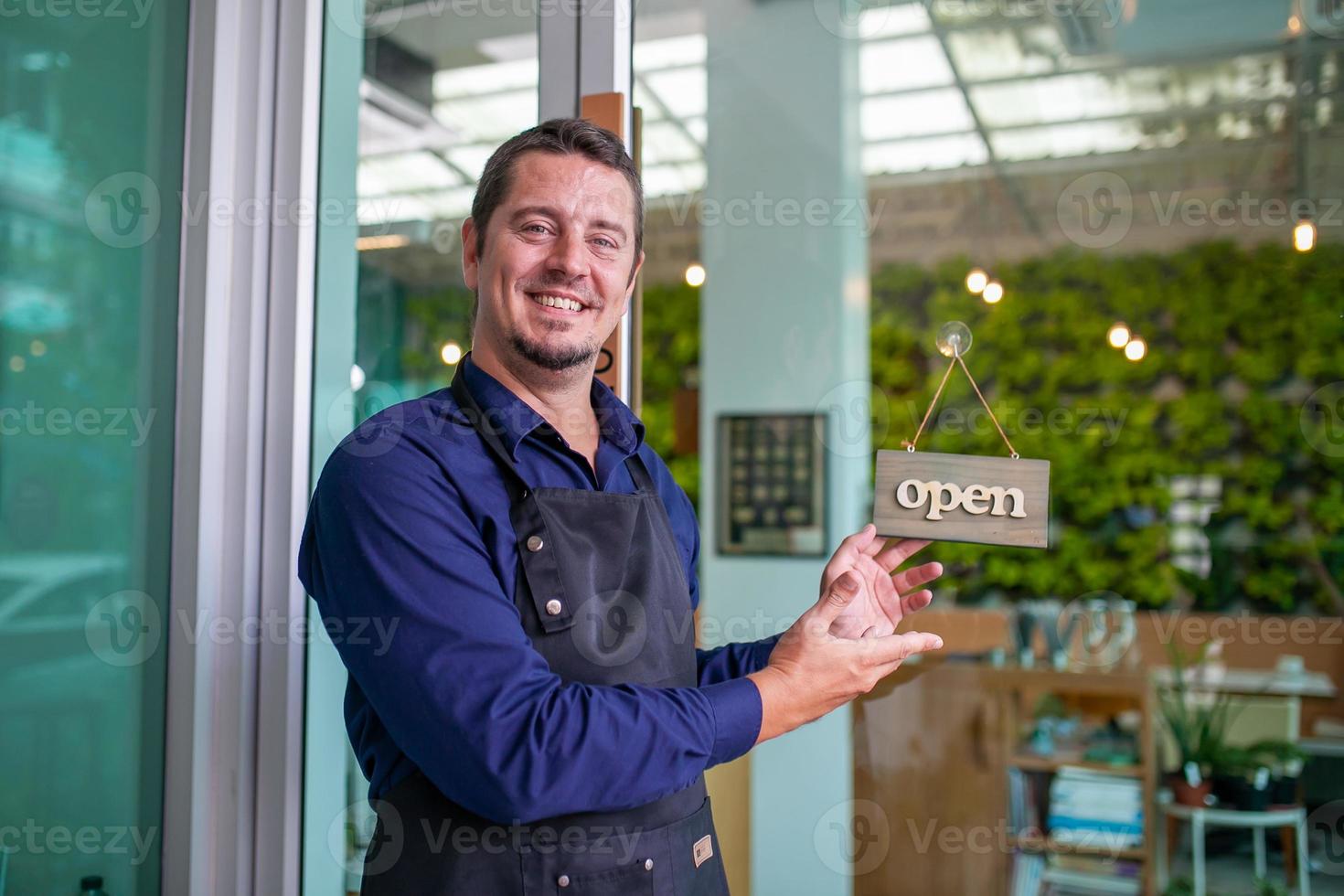 Portrait of smiling owner man standing at his cafe door with open signboard. Coffee owner standing in front of coffee shop to welcome customer and open the coffee shop in morning. photo