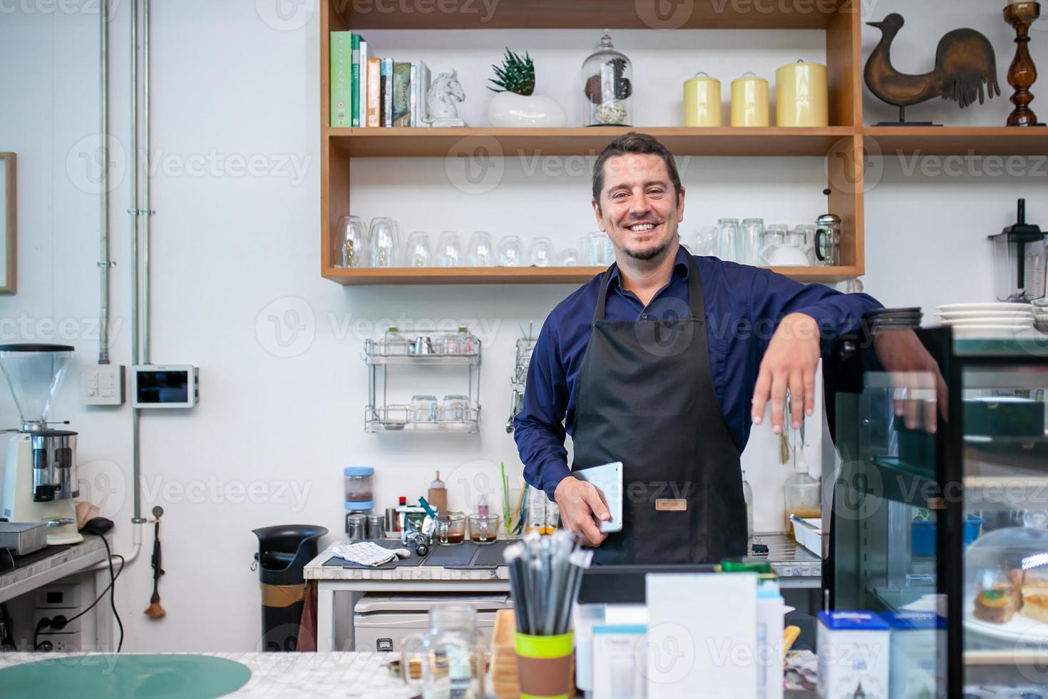 Portrait of smiling owner man standing at his cafe. Coffee owner standing with apron in coffee shop to welcome customer. photo