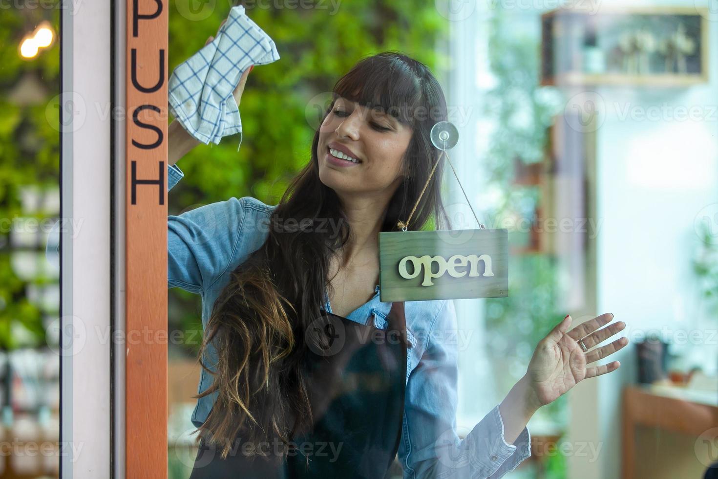 retrato de la dueña sonriente limpiando la puerta de su café con un letrero abierto. dueño del café en la cafetería para dar la bienvenida al cliente y abrir la cafetería por la mañana. foto