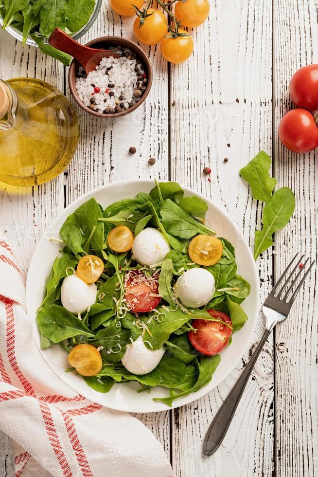 Fresh salad with arugula, cherry tomatoes, mozzarella cheese and hard cheese on white wooden background. Top view photo