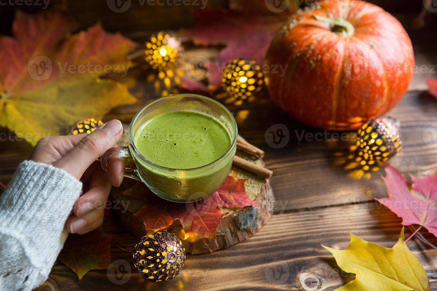 Green Japanese matcha tea with foam in transparent Cup on wooden table in autumn still life. Women's hand with long white sweater sleeves are warmed by drink. Focus on Cup. Warm atmosphere and comfort photo