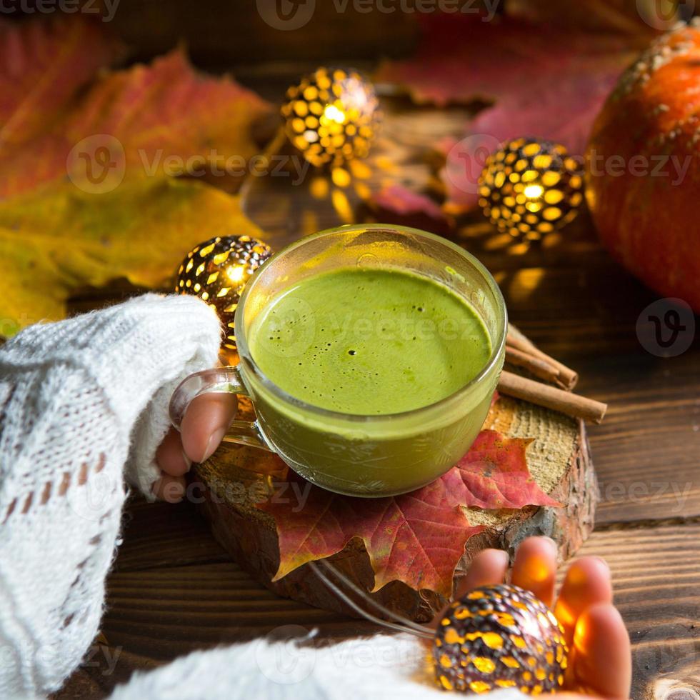 Green Japanese matcha tea with foam in transparent Cup on wooden table in autumn still life. Women's hand with long white sweater sleeves are warmed by drink. Focus on Cup. Warm atmosphere and comfort photo