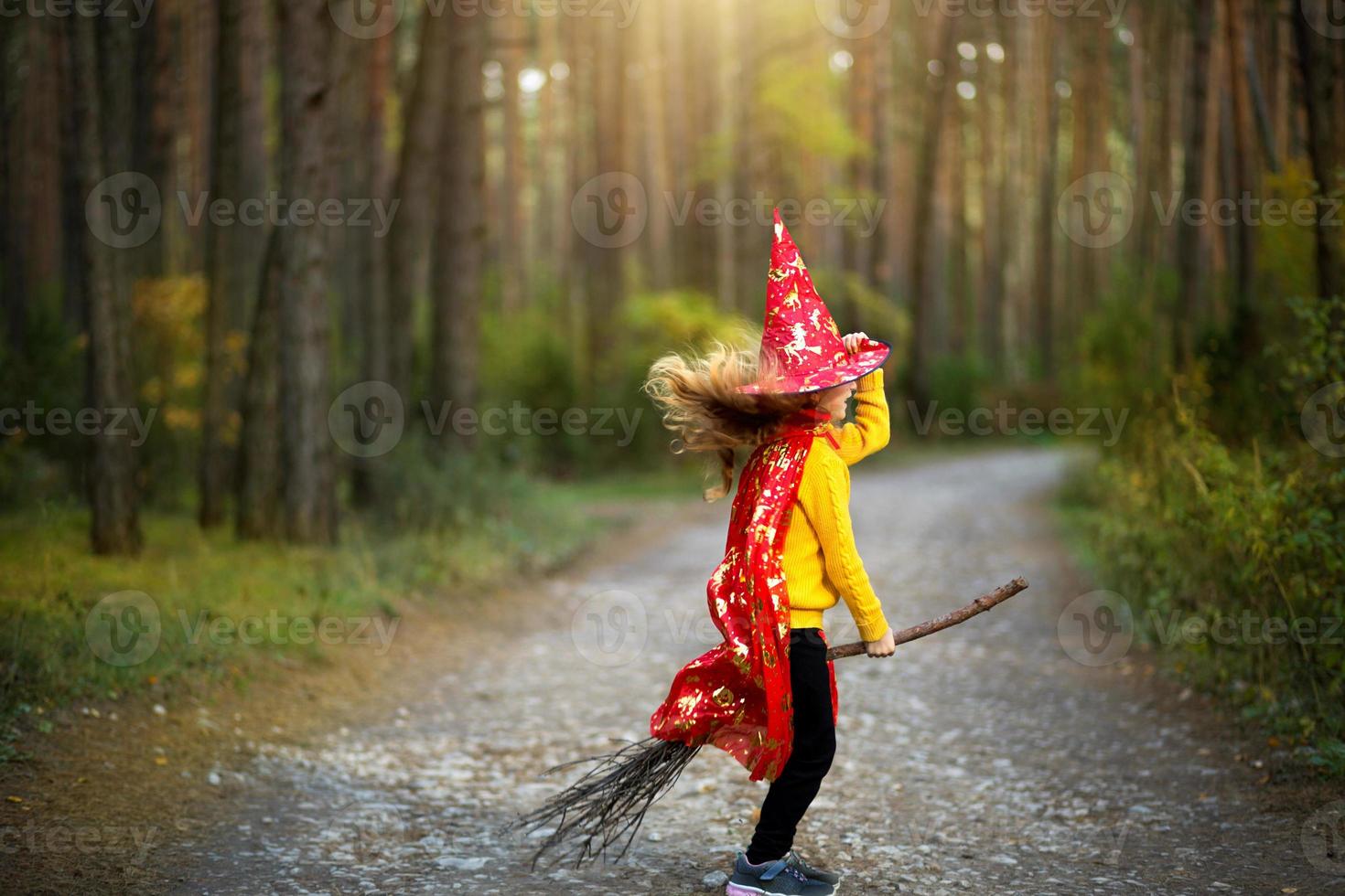 una niña con un disfraz de bruja y un sombrero en una escoba está jugando en el bosque de otoño, yendo a una fiesta de halloween foto