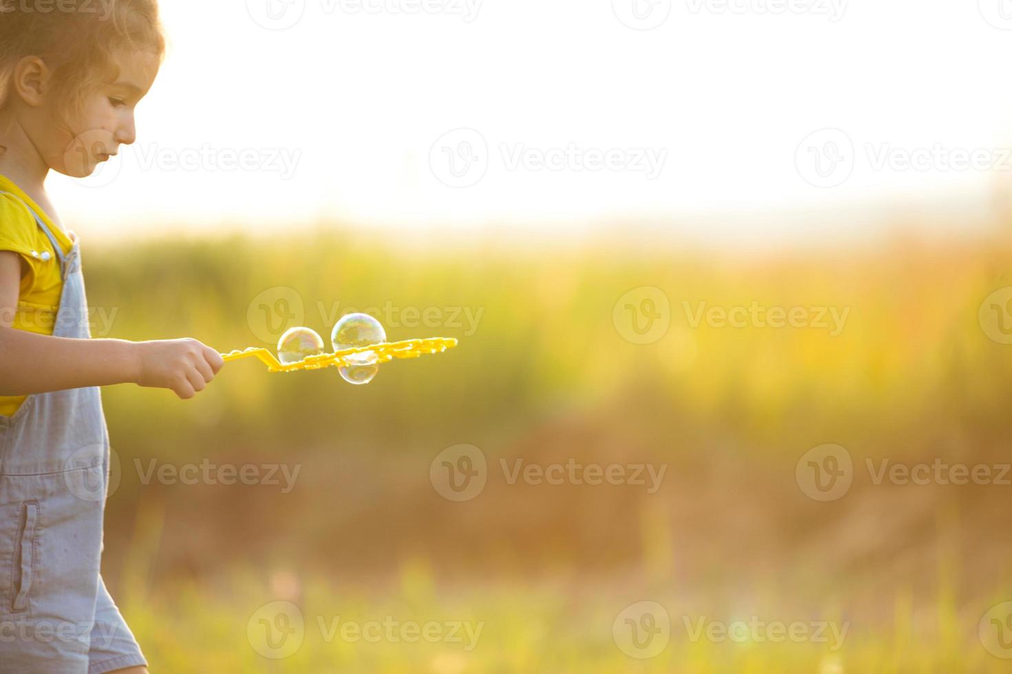 una chica con un traje de mezclilla sopla burbujas de jabón en el verano en un campo al atardecer. día internacional del niño, niño feliz, actividades al aire libre. fondo de verano. estilo de vida saludable y ecológico foto