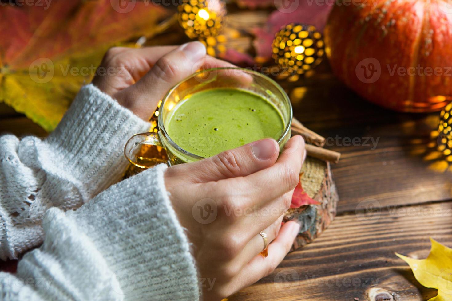 Green Japanese matcha tea with foam in transparent Cup on wooden table in autumn still life. Women's hand with long white sweater sleeves are warmed by drink. Focus on Cup. Warm atmosphere and comfort photo