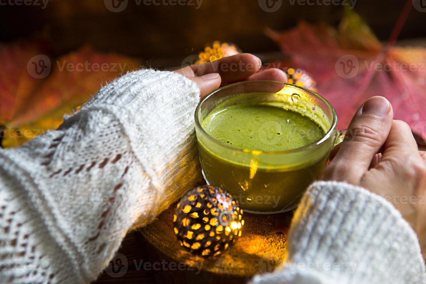 Green Japanese matcha tea with foam in transparent Cup on wooden table in autumn still life. Women's hand with long white sweater sleeves are warmed by drink. Focus on Cup. Warm atmosphere and comfort photo