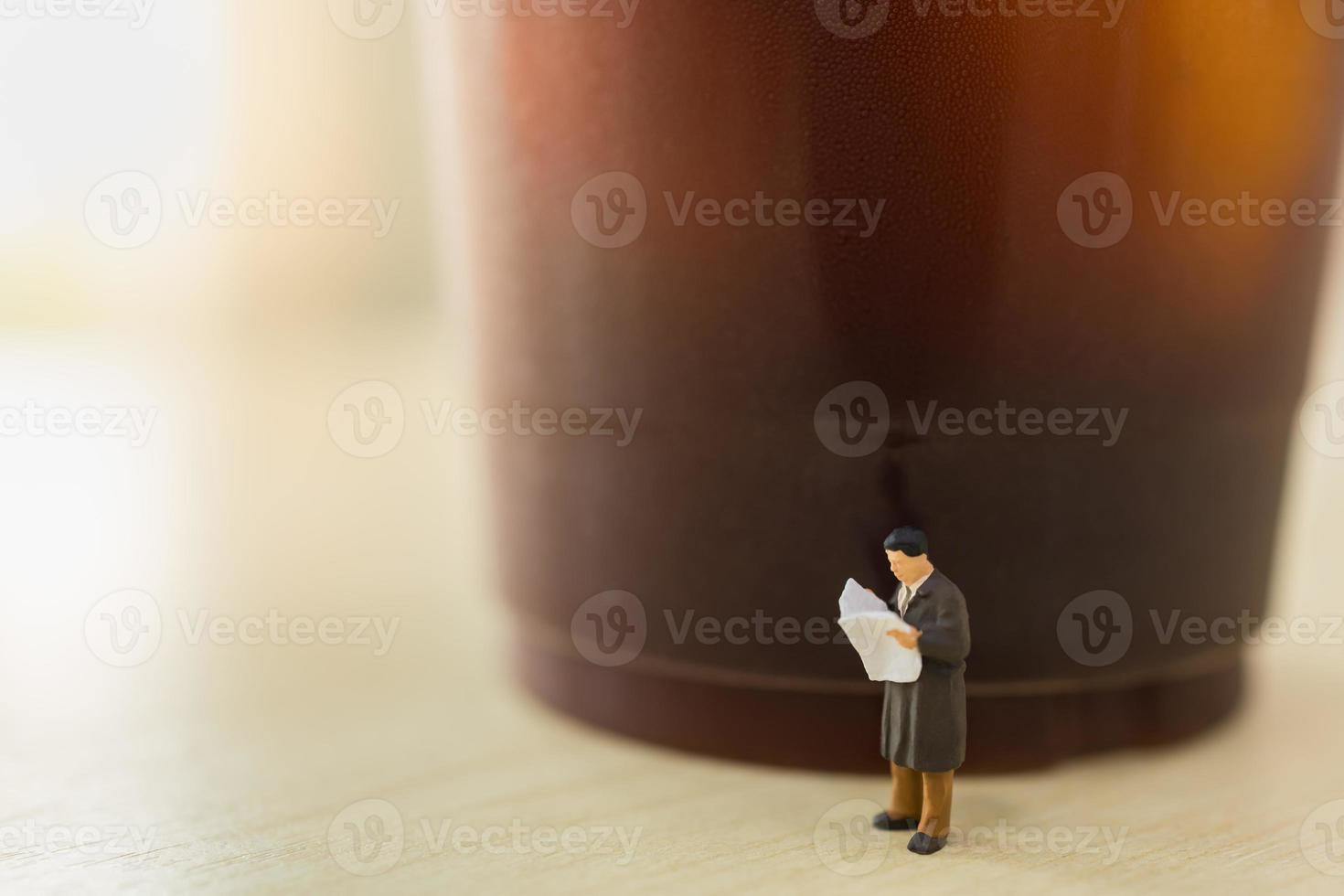 Business, Relaxation Concept. Close up of businessman miniature figure people standing reading newspaper with takeaway plastic cup of iced black coffee on wooden table. photo