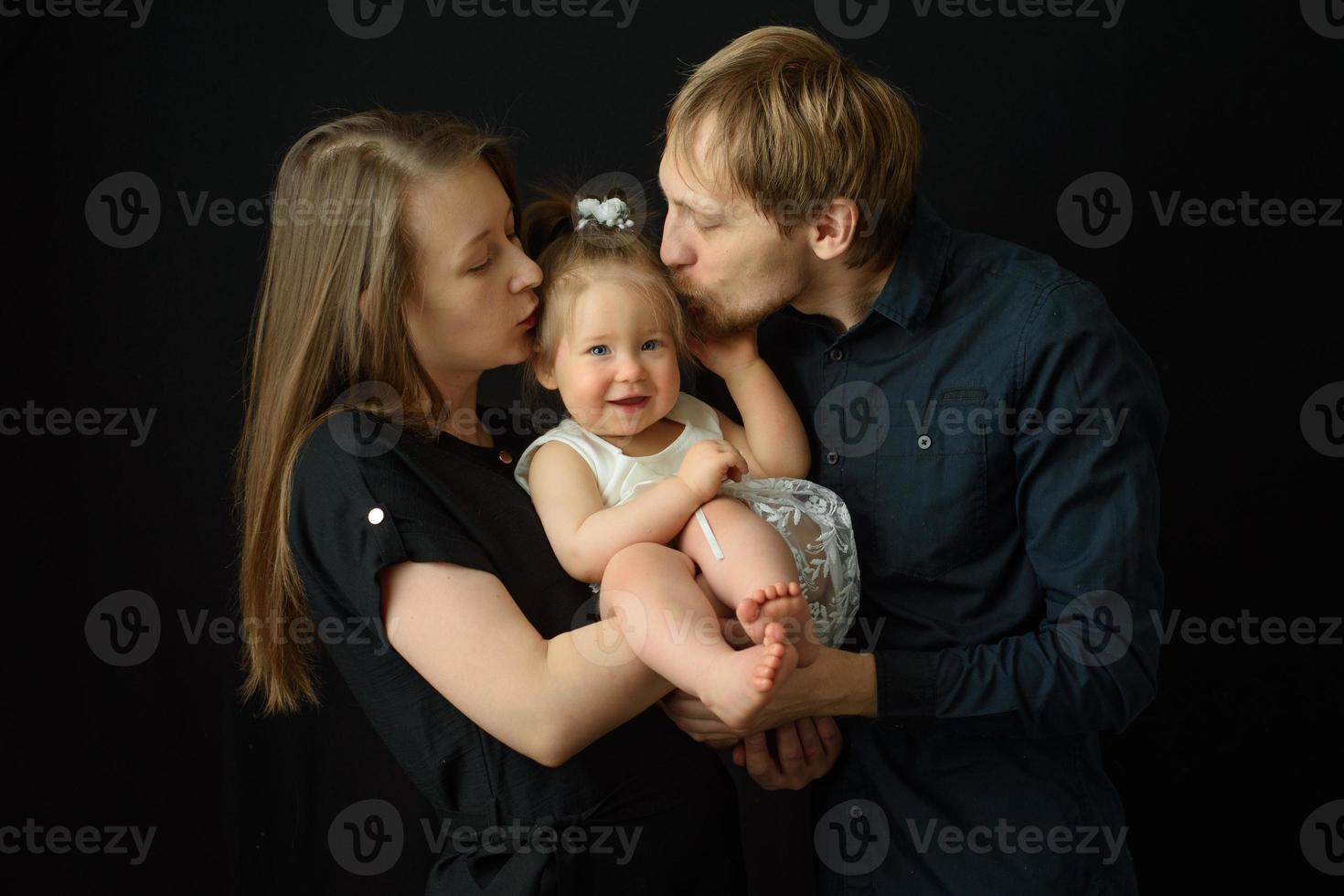 Father and mother kiss their little one year old daughter. Shot on a black background. photo