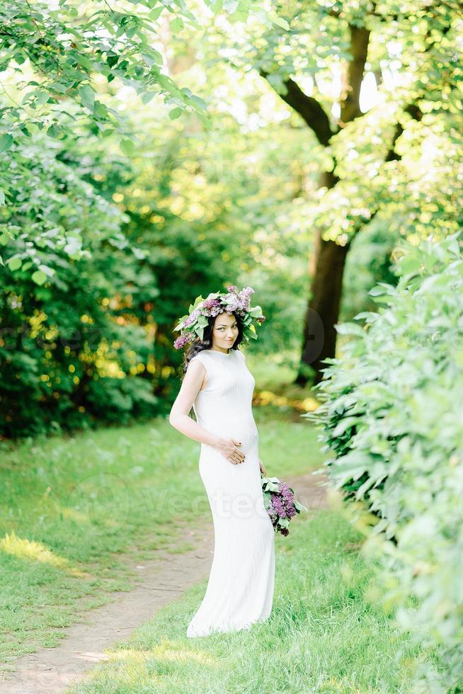 joven embarazada en el jardín floreciente lila de primavera. mirada romántica con sombrero de paja. mujer caucásica con pelo largo y castaño. concepto de nueva vida de la naturaleza y el ser humano. esperando bebe foto