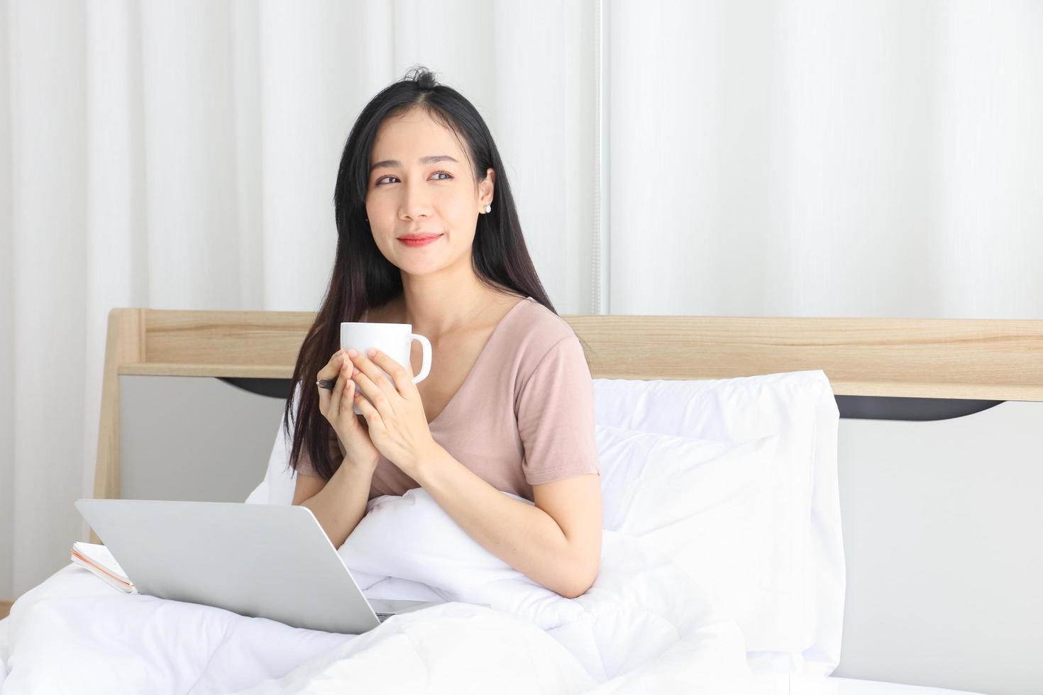 Asian woman working online from home with laptop while drinking coffee on her bed for new normal life during quarantine period from covid-19 photo