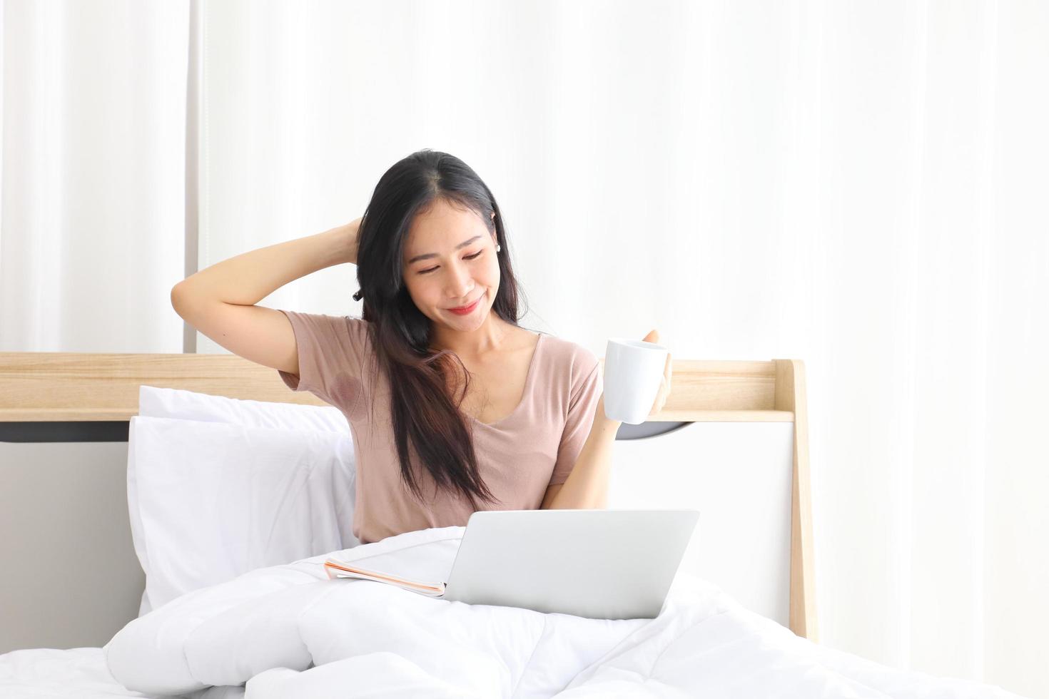 jovencita asiática que trabaja desde casa en su cama usando una computadora portátil para acceder a su trabajo en línea con espacio para copiar foto