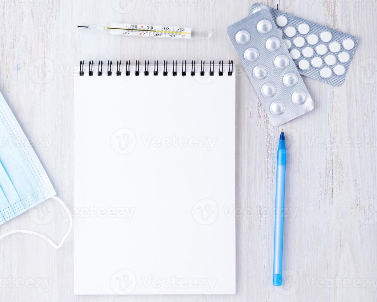 Top view on the table in the doctor's office. Open notepad with a blank page, medicine, pen, mask, thermometer. Write a treatment plan photo