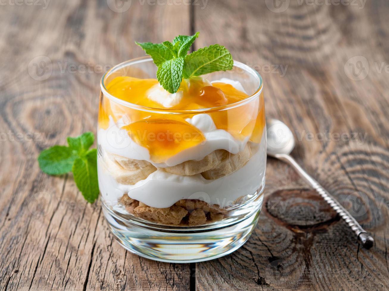 Healthy layered dessert with yogurt, banana, mango, cookie on wooden background, side view. photo