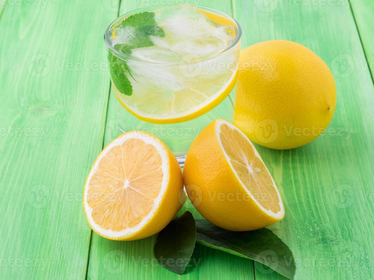 Lemonade in a glass, a lemon half, fresh leaves on the green table. A refreshing cold drink of water with ice, mint and slices of lemon photo