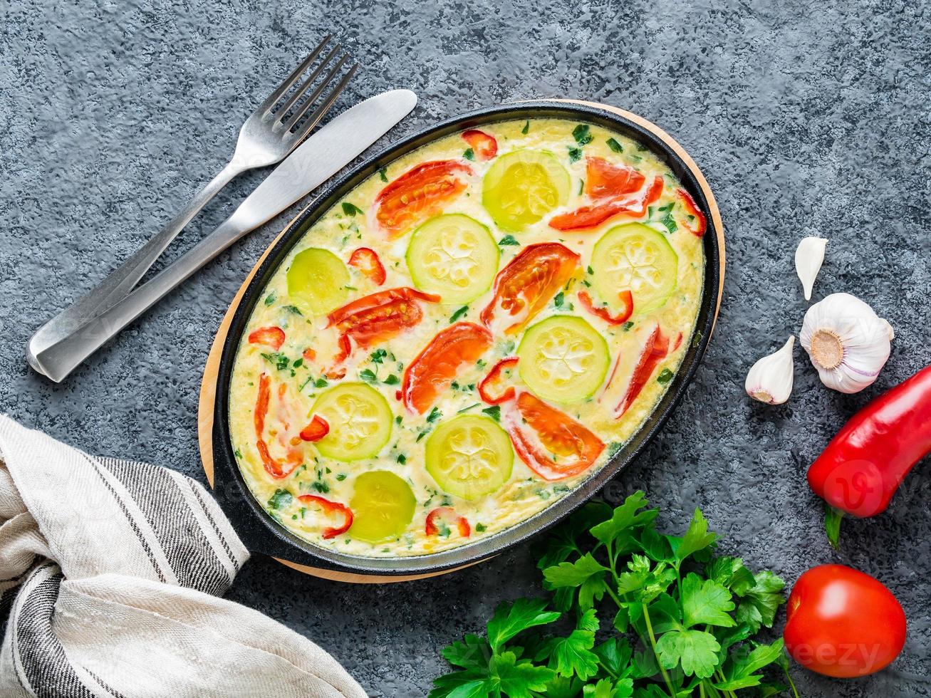 sartén con frittata italiana tradicional sobre mesa de piedra gris oscuro, tenedores, perejil, pimienta, tomates. huevos revueltos foto