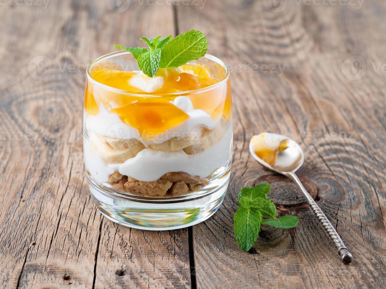 Healthy layered dessert with yogurt, banana, mango jam, cookie on wooden background, side view photo