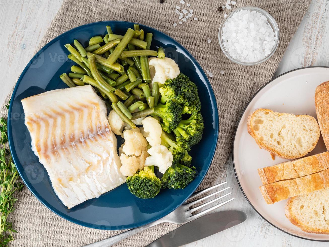 Baked sea fish cod fillet with vegetables on blue plate, bread, gray napkin, wooden background, top view. Proper diet photo