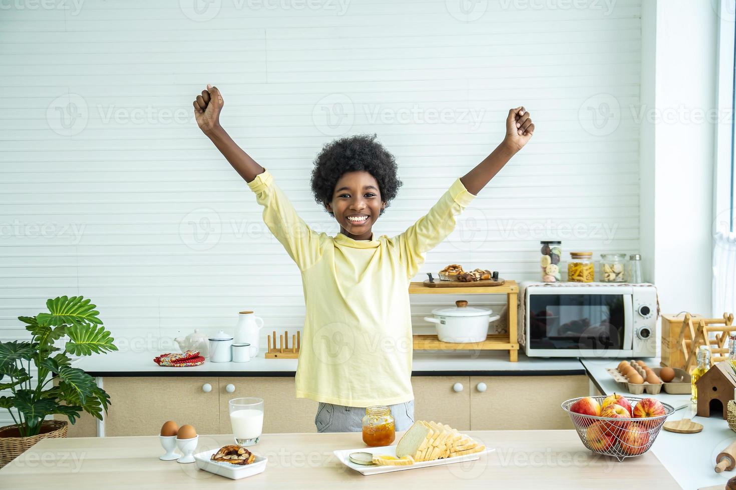 adorable y feliz niño pequeño levantando las manos, niño disfrutando de su delicioso desayuno en la cocina en casa por la mañana foto