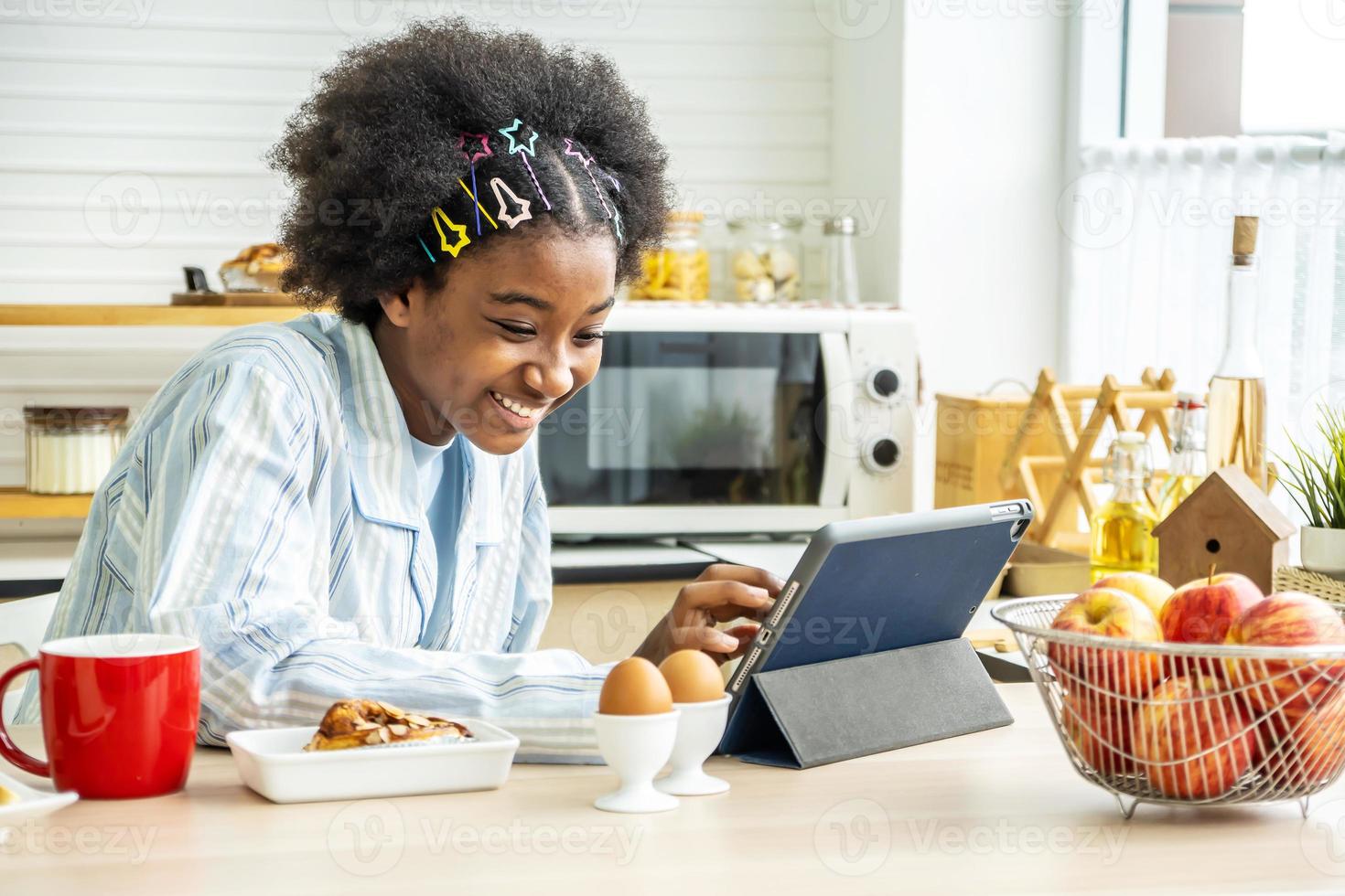 Young woman african american in the kitchen while having breakfast and sipping tea or milk in the cup send a message or calls with the tablet and smiles, Concept of social network, message, technology photo