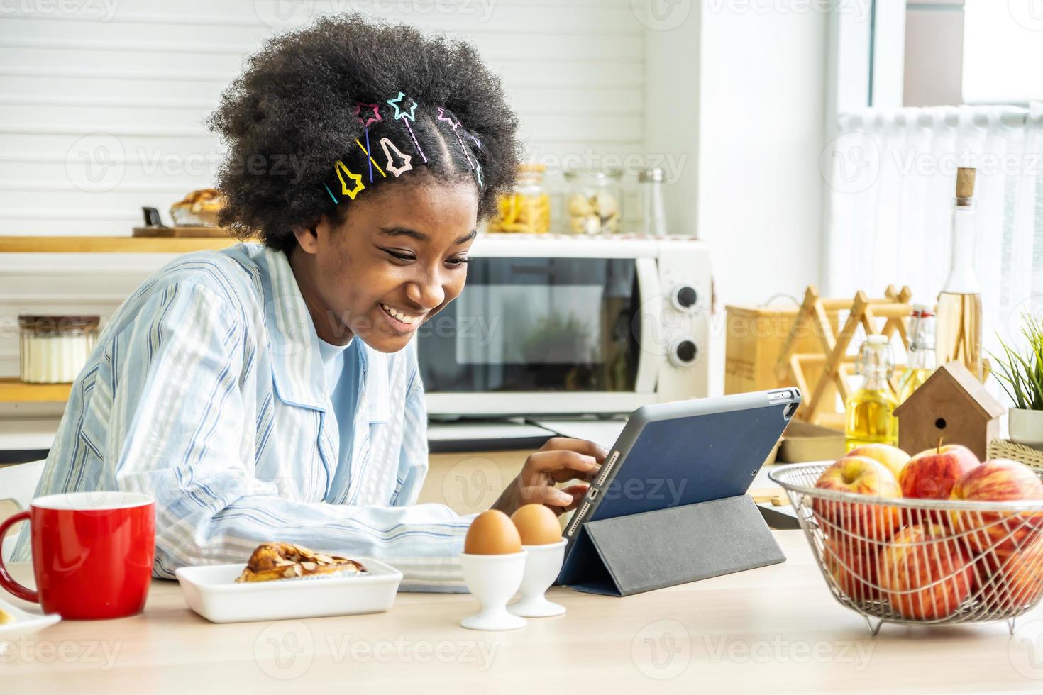 mujer joven afroamericana en la cocina mientras desayuna y toma té o leche en la taza envía un mensaje o llama con la tableta y sonrisas, concepto de red social, mensaje, tecnología foto