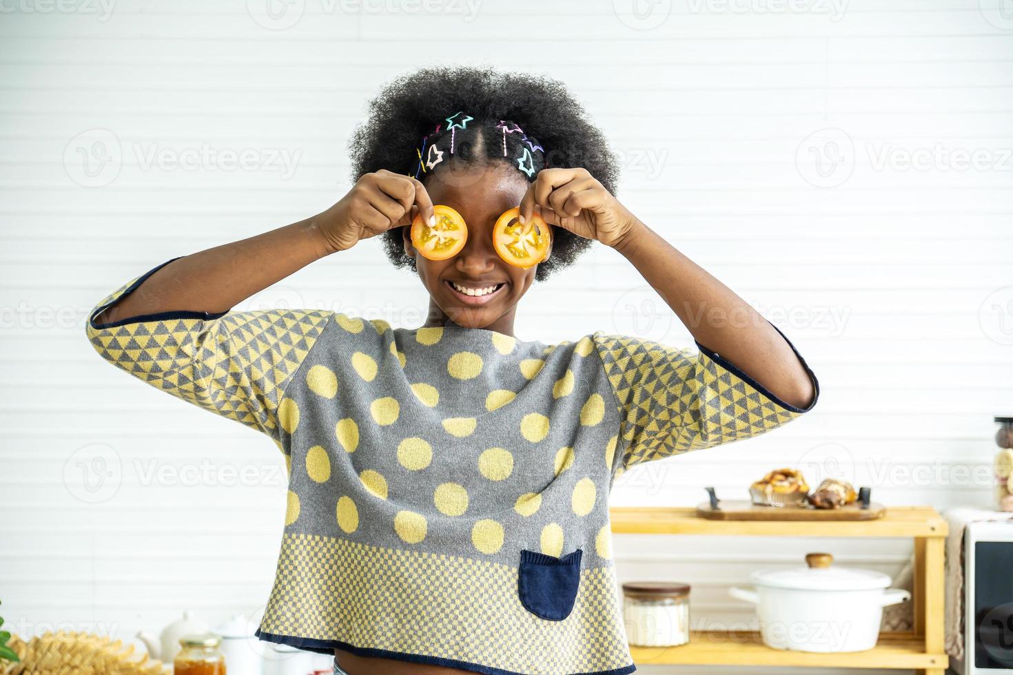 African american happy girl having fun with food vegetables at kitchen holds tomatoes before his eyes like in glasses, Teaching child to healthy and varied vegitarian food photo