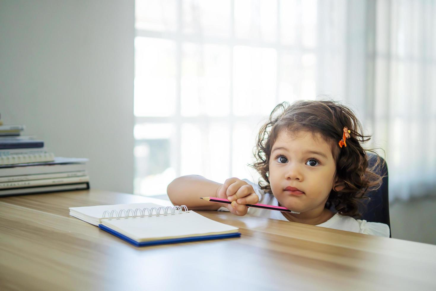 linda niña sentada en el escritorio en casa haciendo deberes, leyendo, escribiendo y pintando. los niños pintan. los niños dibujan niño en edad preescolar con libros en casa. los preescolares aprenden a escribir y leer. niño creativo foto