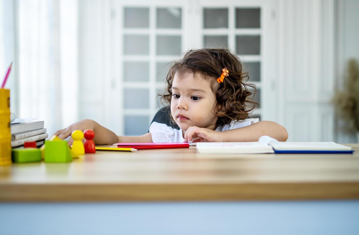 Cute little girl sit at desk at home doing homework, reading, writing and painting. Children paint. Kids draw. Preschooler with books at home. Preschoolers learn to write and read. Creative toddler photo