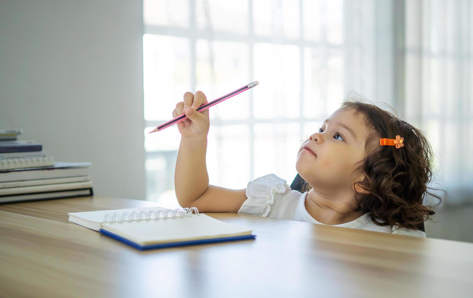 linda niña sentada en el escritorio en casa haciendo deberes, leyendo, escribiendo y pintando. los niños pintan. los niños dibujan niño en edad preescolar con libros en casa. los preescolares aprenden a escribir y leer. niño creativo foto