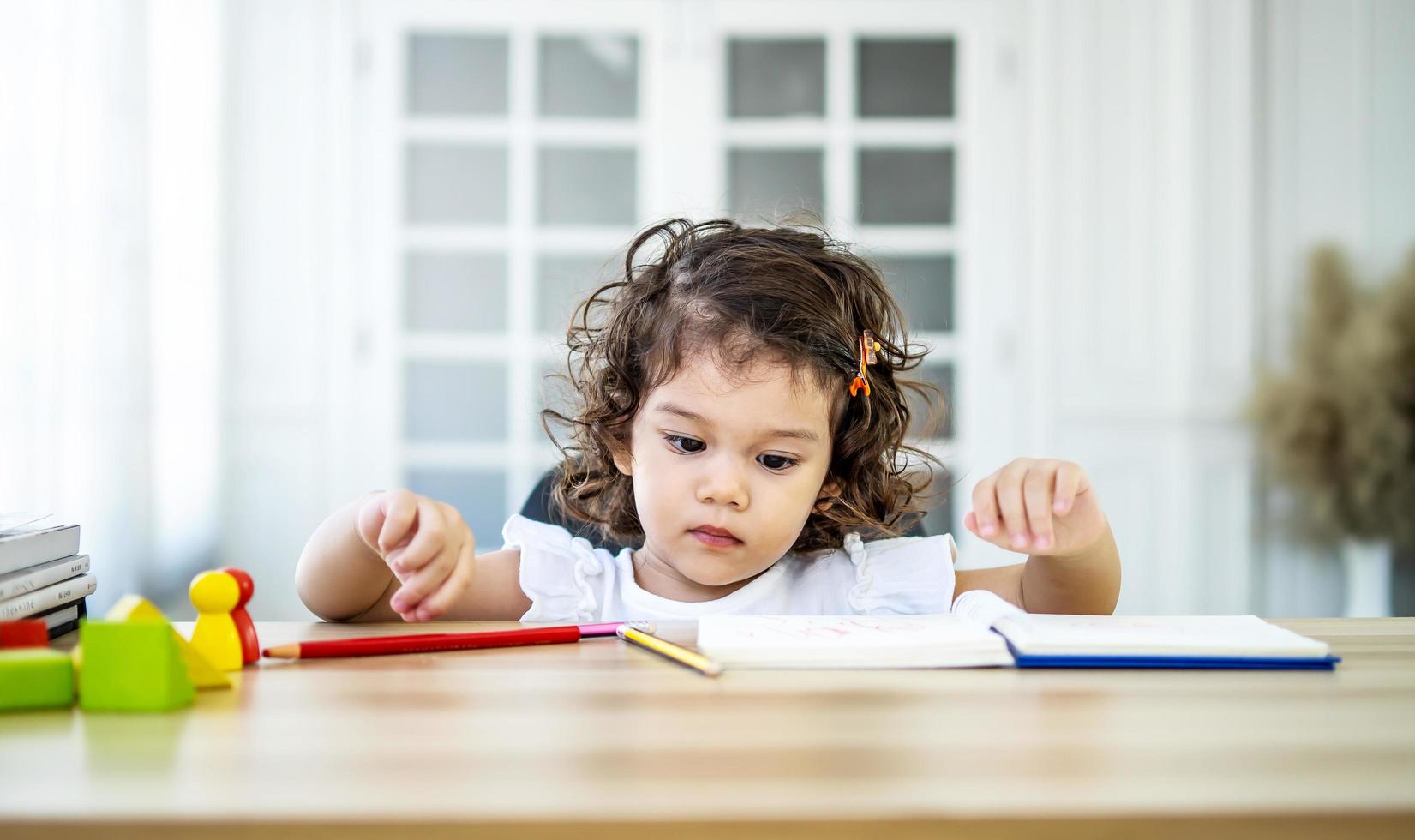 linda niña sentada en el escritorio en casa haciendo deberes, leyendo, escribiendo y pintando. los niños pintan. los niños dibujan niño en edad preescolar con libros en casa. los preescolares aprenden a escribir y leer. niño creativo foto