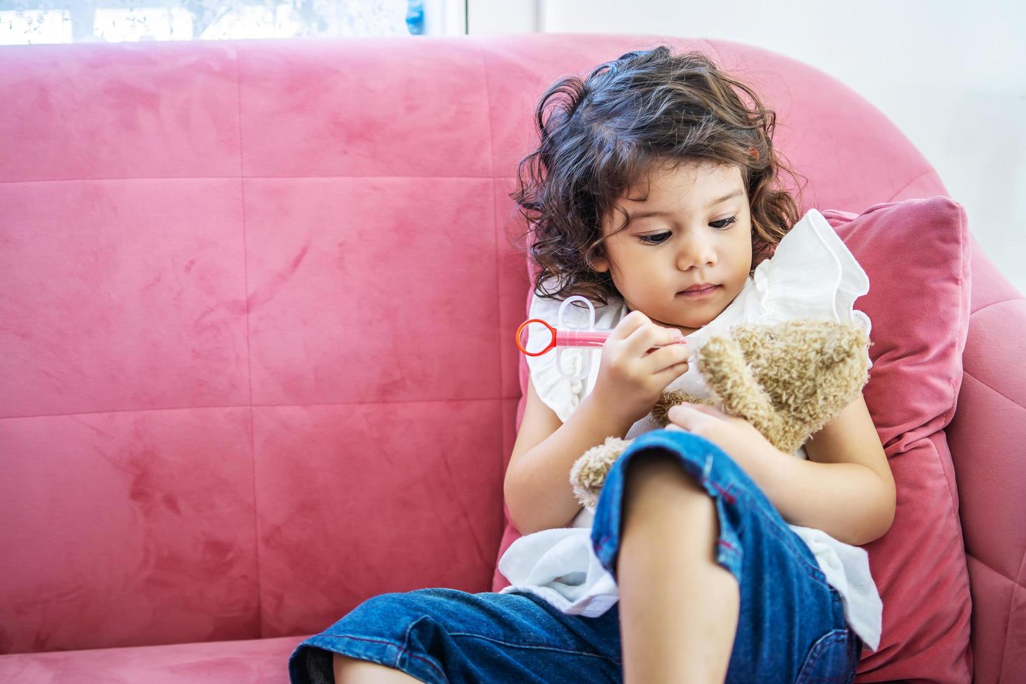 una niña linda disfruta jugando con jeringas de inyección de juguete en el oso de peluche en el sofá de la casa, tiempo de educación en casa foto
