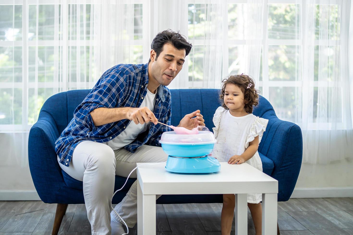 Hand father rolling cotton candy in candy floss machine and eat cotton candy together with daughter on sofa at new house photo