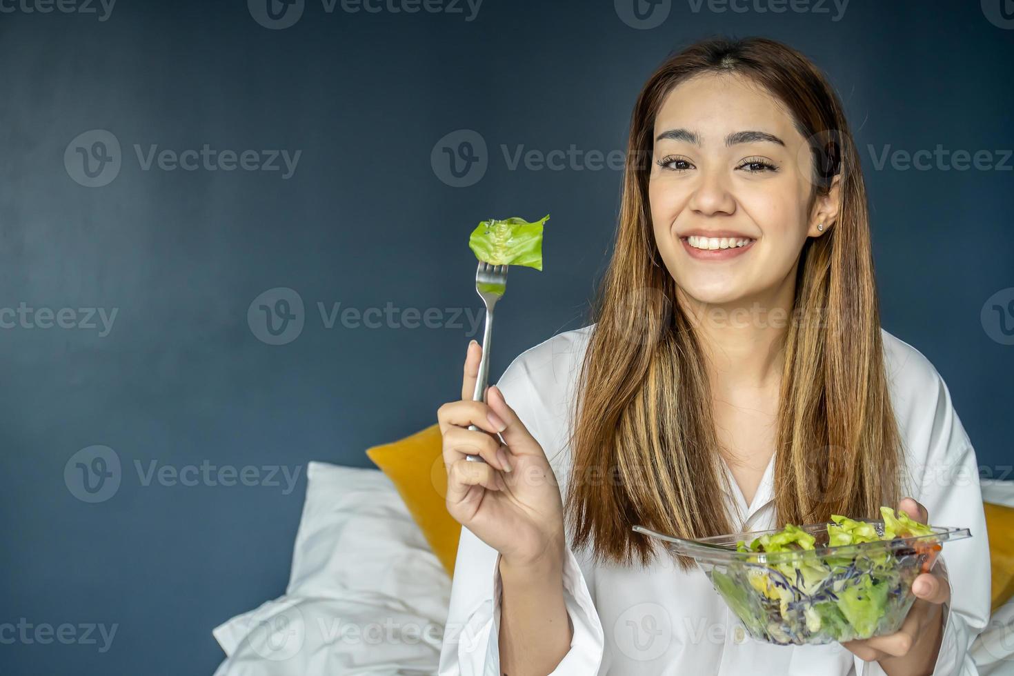 Gorgeous young woman sitting on bed eating vegetable salad, Balanced raw food diet. Positive girl having vegan lunch, taking care of her wellbeing photo