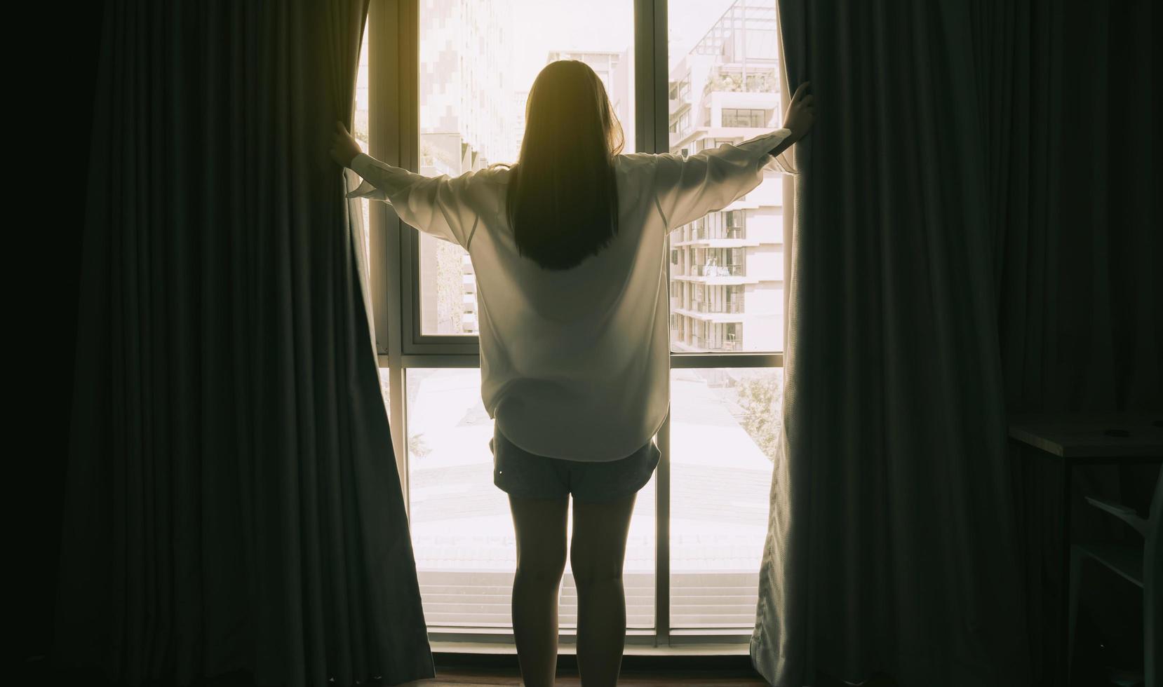 Overjoyed young beautiful woman standing by bedroom and opening curtains look in window distance meet welcome new day. Smiling female feel excited about life career opportunities or perspectives. photo