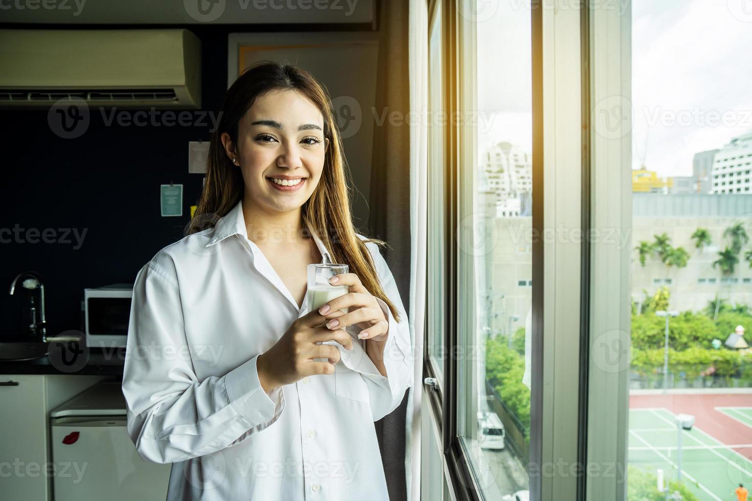 Young asian woman looking out window enjoying fresh new day feeling rested and drinking glass of milk at hotel. photo