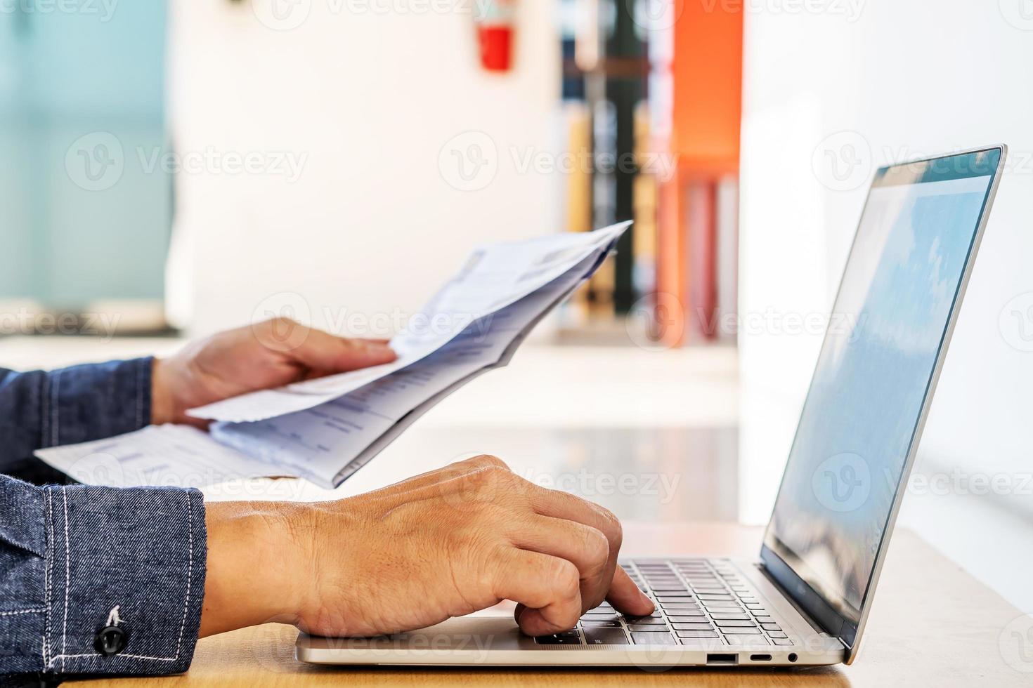 hombre de negocios revisando un informe o documento escrito mientras se sienta a trabajar en papeleo en una computadora portátil, pagando facturas en línea en una computadora portátil foto