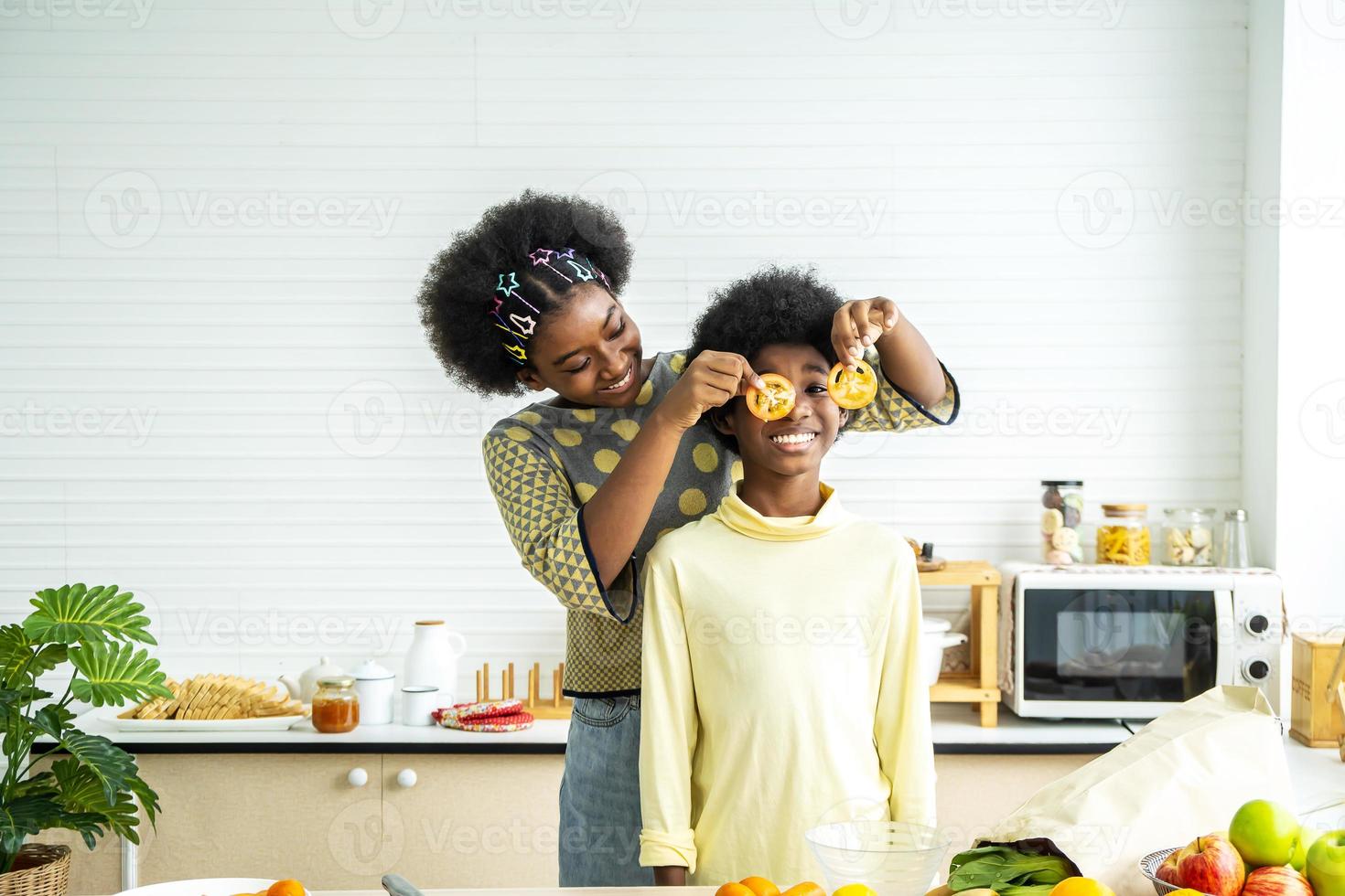 dos niños afroamericanos disfrutan y se divierten felices con verduras en la cocina sostienen tomates ante sus ojos como en vasos, enseñando a los niños comida vegetariana sana y variada foto