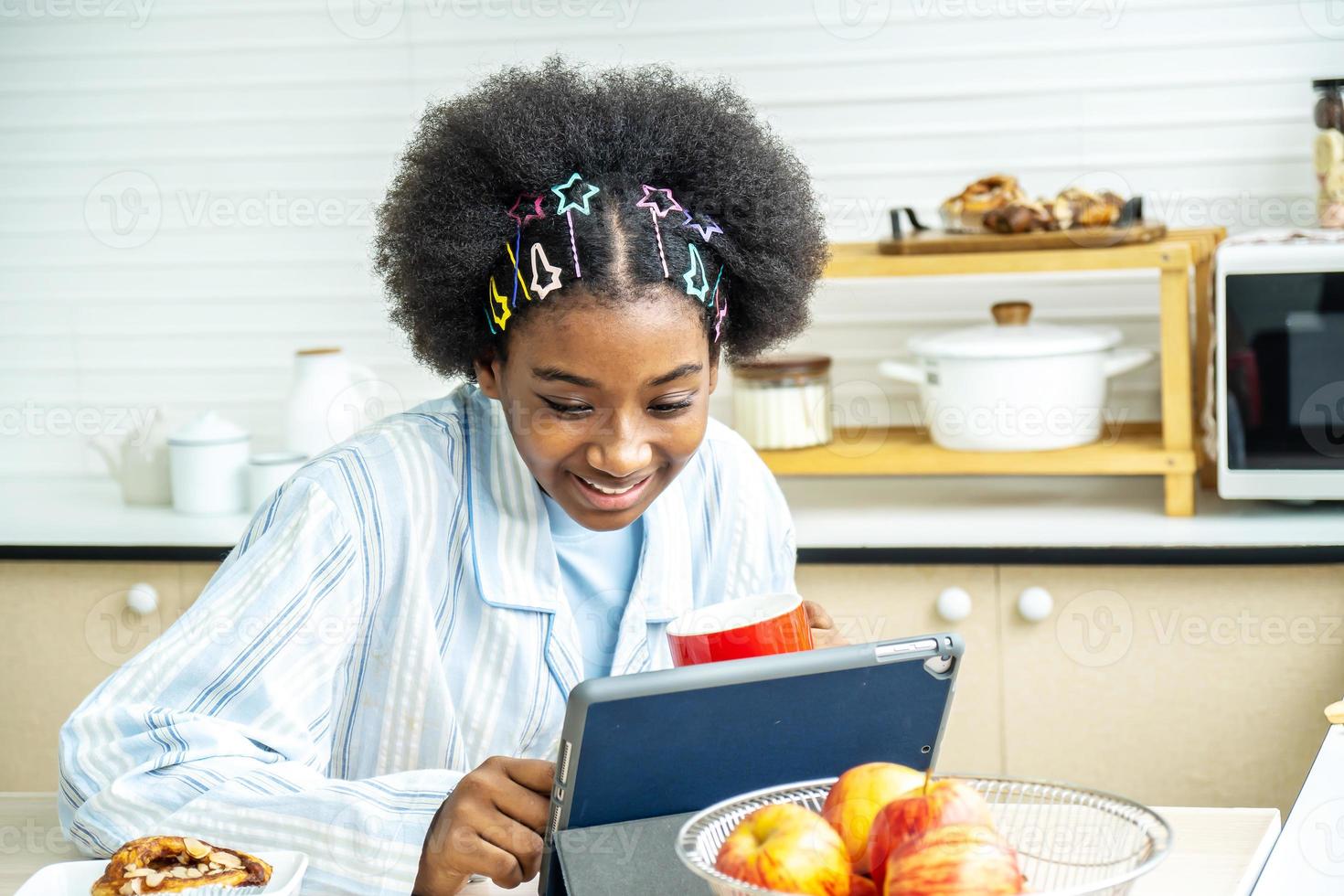 Young woman african american in the kitchen while having breakfast and sipping tea or milk in the cup send a message or calls with the tablet and smiles, Concept of social network, message, technology photo