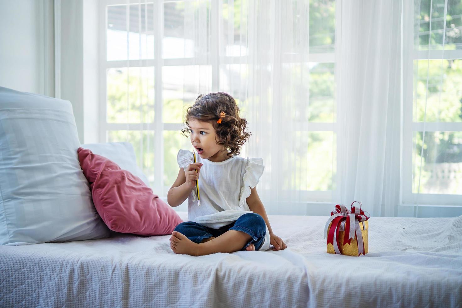 take congratulations on your birthday. A little girl in curlers sits on the bed in the bedroom playing with a pencil. Party preparation. Funny child with a pencil. photo